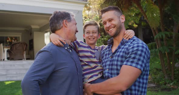 Portrait of happy caucasian grandfather, grandson and father embracing in garden and smiling