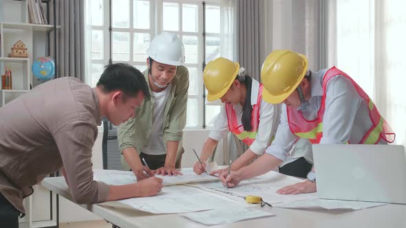 Three Asian Engineers With Helmets Helping A Man Drawing Building Construction At The Office