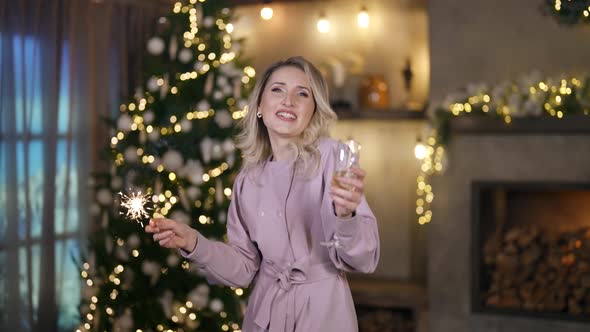 a Blonde in a Festive Dress Sings and Dances with a Glass and a Sparkler in Hands Against the