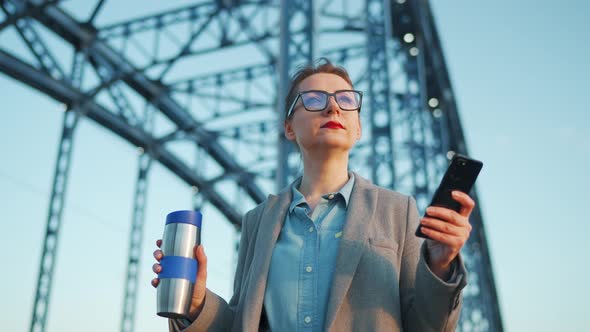 Woman in a Coat Walking Around the City in the Early Morning Drinking Coffee and Using Smartphone