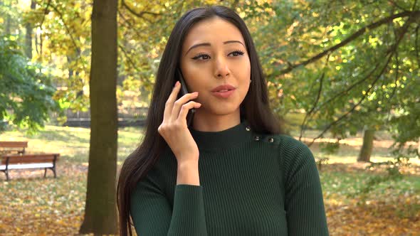 A Young Asian Woman Talks on A Smartphone in A Park