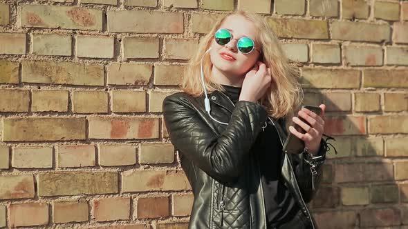 Stylish Young Blonde in Sunglasses and Leather Jacket Listening to Music on Bluetooth Headphones in