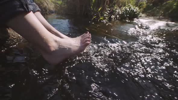 Woman Relaxes By River Sitting on Edge of Rock Swing One's Feet on Water Flow