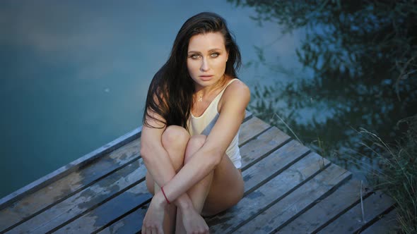 A Beautiful Girl at Dusk Sitting on a Wooden Pier Near the Water