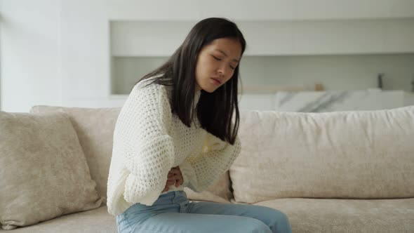 An Asian Woman in a White Blouse is Sitting on the Couch and Feels a Strong Abdominal Pain Which is
