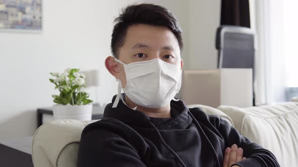 A Young Asian Man in a Face Mask Looks Seriously at the Camera As He Sits on a Couch at Home