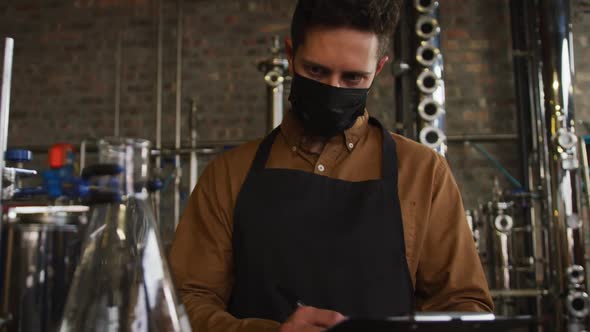 Portrait of caucasian man working at gin distillery wearing face mask and apron looking to camera