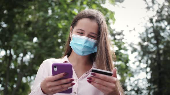 Happy girl wearing medical mask outdoor holding credit card and using smart mobile payments.