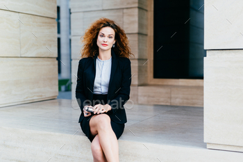 Attractive young woman wearing black formal suit, sitting crossed legs with electronic device.