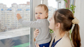 Young mother holding her baby son trying to open window on high floor. Baby in danger. Child safety - PhotoDune Item for Sale