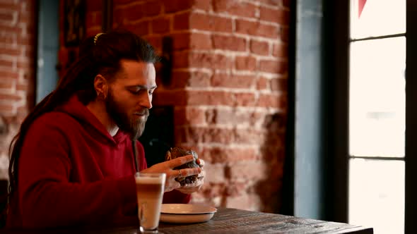 A Man with Long Hair and Beard Greedily Eats a Burger in a Cafe