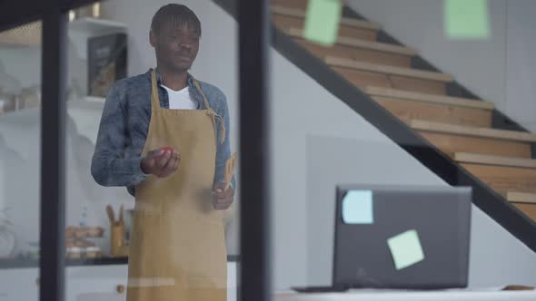 Shooting Through Glass of Man in Apron Standing in Kitchen Talking at Laptop Web Camera