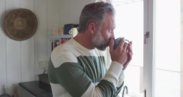 Caucasian mature man looking through the window and drinking coffee
