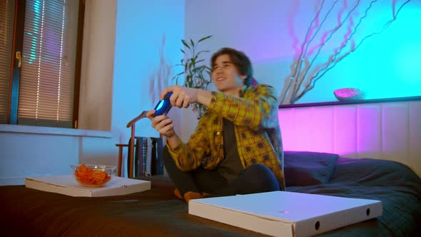 Young Excited Man Sitting on the Bed Enthusiastically Playing Video Game and Eating Chips