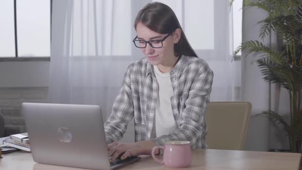 Cute Caucasian Brunette Girl in Elegant Eyeglasses Typing on Laptop Keyboard. Confident Smiling