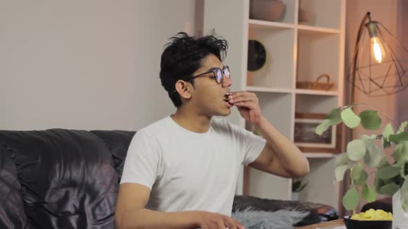 Young man with glasses eats potato chips and falls on couch, relaxing time