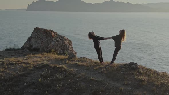 Two Women in Sportswear and Barefoot Doing Yoga Exercises Holding Each Other's Hands Against the