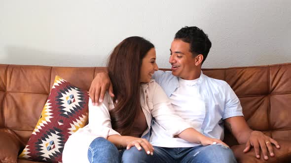 Young couple at home sitting on the sofa looking each other with love and smiling - concept of love