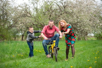 ple in spring garden. Family having fun, male riding on children’s bicycle, woman and a boy of his support
