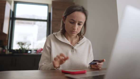 Irritated Young Caucasian Girl Sitting with Computer and Credit Card on Couch in Kitchen Room Can