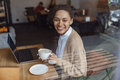 Young woman holding a cup with cappuccino, enjoying remote work on laptop in coffee shop - PhotoDune Item for Sale