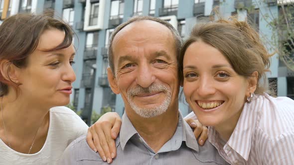 Family Portrait of Senior Father and Two Adult Daughters Kissing Him Outdoors on Modern Building of