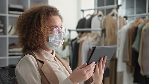 Portrait of a Young Woman in a Medical Mask and Glasses Uses a Tablet for Online Sales in an