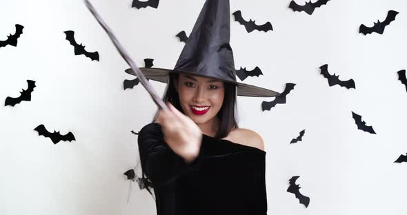 Playful Asian Lady in Sorceress Costume Making Halloween Magic, Gesturing at Camera with Wand