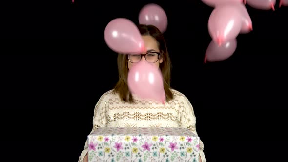 A Young Woman Is Standing with a Gift, and Balloons Are Falling on Her. Smiling Woman Holding a Gift