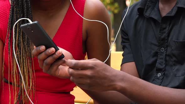 A Black Man and a Black Woman Sit on a Bench in a Park and Listen To Music on a Smartphone - Closeup