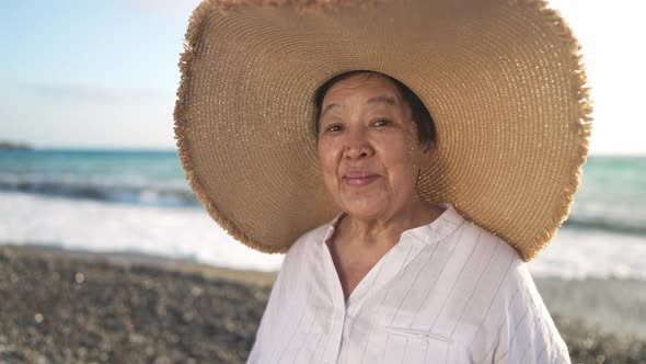 Happy Asian Old Woman in Straw Hat Looking at Camera Smiling Posing on Picturesque Mediterranean Sea