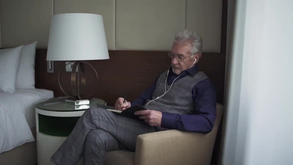 Portrait of an Grayhaired Man Sitting in a Hotel Room and Writing Down Thoughts in a Notebook