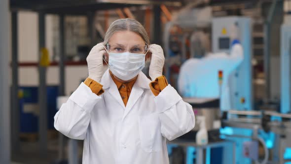 Portrait of Mature Woman Scientist in Robe and Protective Glasses Putting on Safety Mask Standing