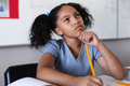 Thoughtful biracial elementary girl with hand on chin looking up while studying at desk in classroom - PhotoDune Item for Sale
