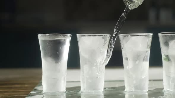 the bartender pours vodka from a bottle into several icy glasses