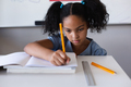 Biracial elementary schoolgirl writing on book at desk while studying in classroom - PhotoDune Item for Sale