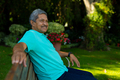 Smiling biracial senior man looking away while sitting on bench against plants in park - PhotoDune Item for Sale