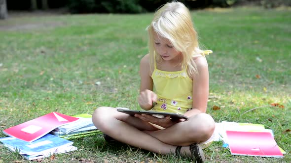 Little Smart Girl Sits in the Park and Searchs Something on the Tablet - Surrounded By Workbooks