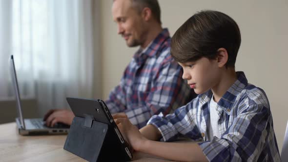 Male Kid Playing Game on Tablet While Father Working on Laptop at Home, Gadget