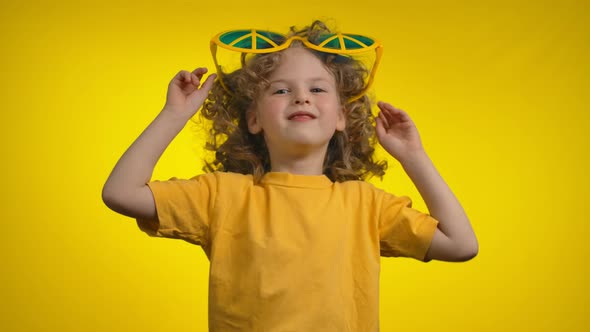 Curly Little Girl in Funny Hippie Sunglasses is Dancing on the Yellow Background