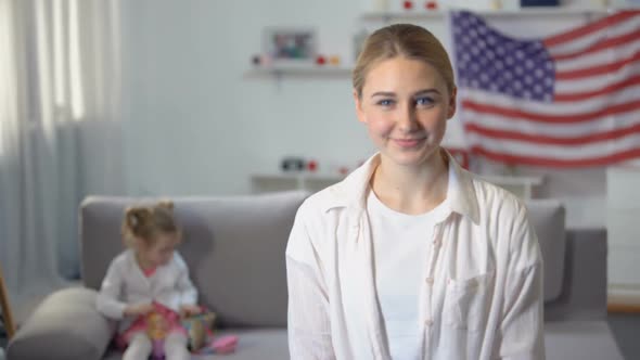 Joyful Patriot Woman Looking at Camera Daughter Playing on Sofa Against US Flag