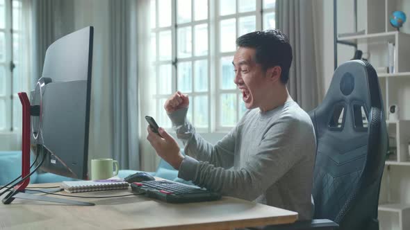 Asian Man Looking At Mobile Phone And Being Happy Winning The Project Bid While Use Computer at Home