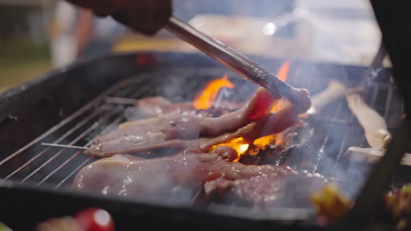 Close up of barbeque grilling meat on bbq grid burning fire with smoke at dark night in camp.