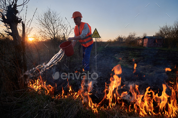 evening sky on background. Male environmentalist pouring water on burning dry grass near yellow triangle with skull and crossbones warning sign.