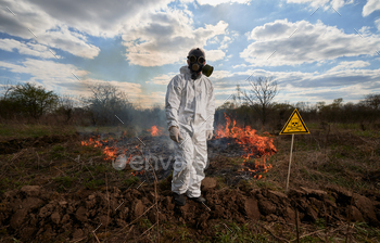 Man in gas mask near burning grass with smoke and yellow triangle with skull and crossbones warning sign. Natural disaster concept.
