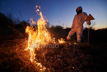 re at night. Man in suit and gas mask near burning grass with smoke, holding warning sign with skull and crossbones. Natural disaster concept.