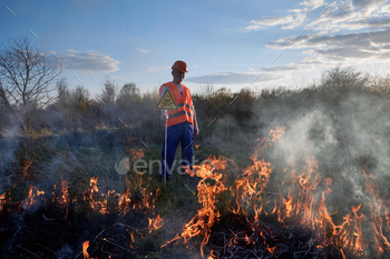 n evening. Man in orange vest and helmet near burning grass with smoke, holding warning sign with skull and crossbones. Natural disaster concept.