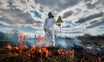 in protective suit and gas mask near burning grass with smoke, holding warning sign with skull and crossbones. Natural disaster concept.