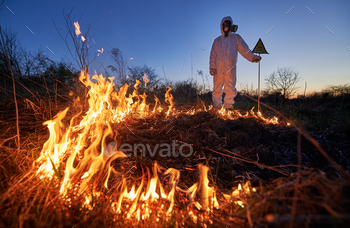 re at night. Man in suit and gas mask near burning grass with smoke, holding warning sign with skull and crossbones. Natural disaster concept.