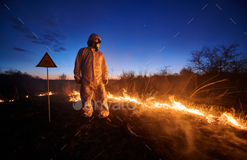 n in protective radiation suit and gas mask near burning grass with smoke and warning sign with skull and crossbones. Natural disaster concept.